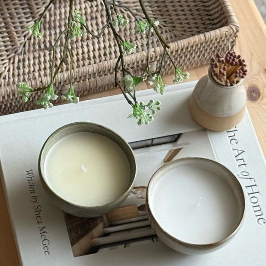 Two small candles in ceramic holders on a magazine with a vase of sticks and branches in the background.