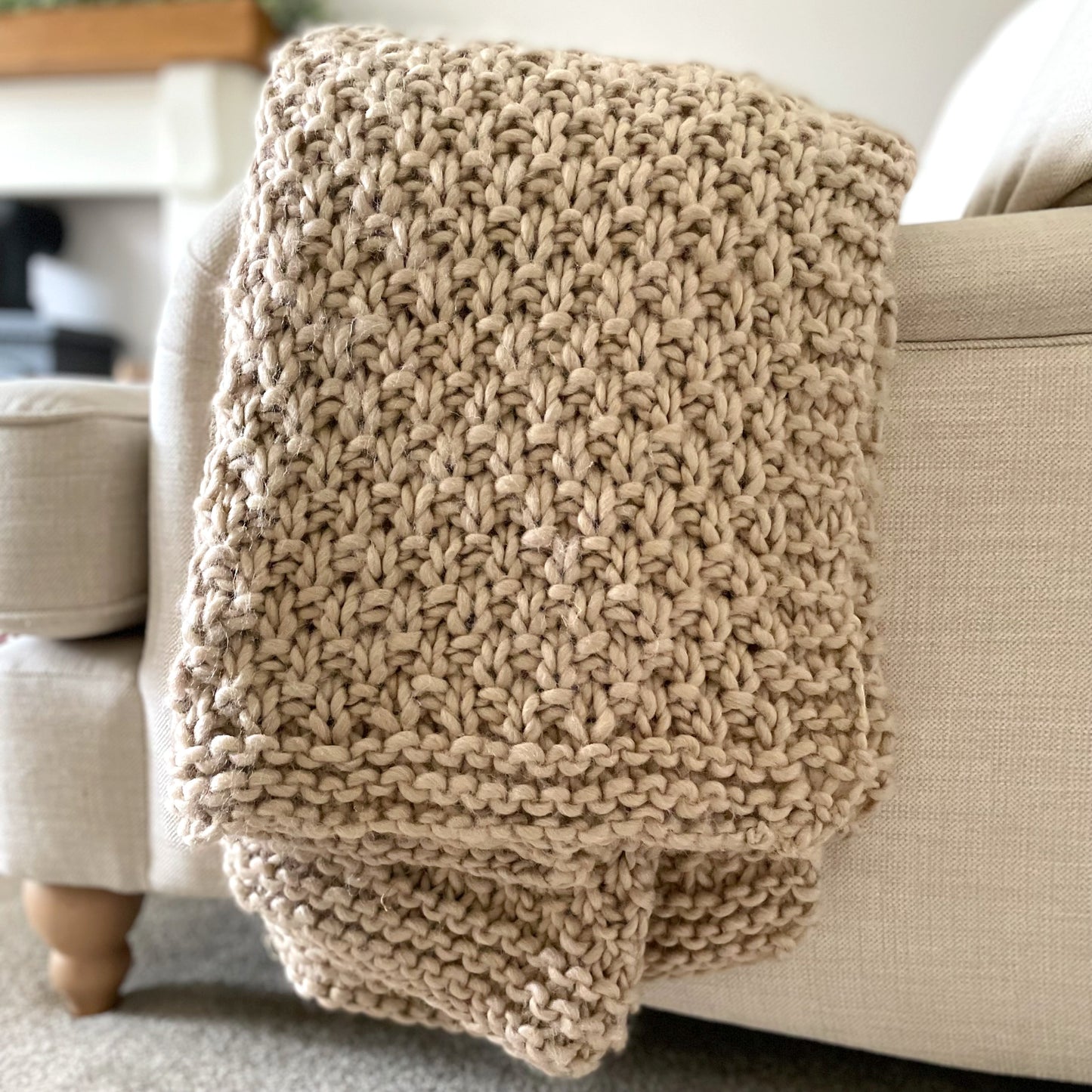 Chunky moss knitted oatmeal throw draped over a cream armchair in a cosy living room.