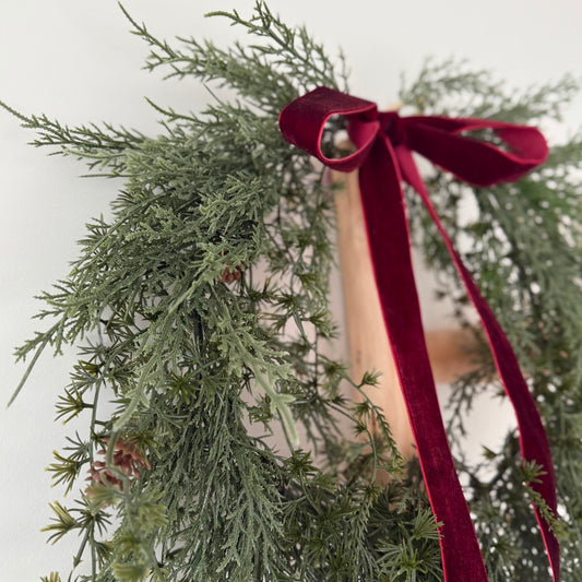 Green faux foliage wreath with mini pinecones with a red ribbon against a light background.
