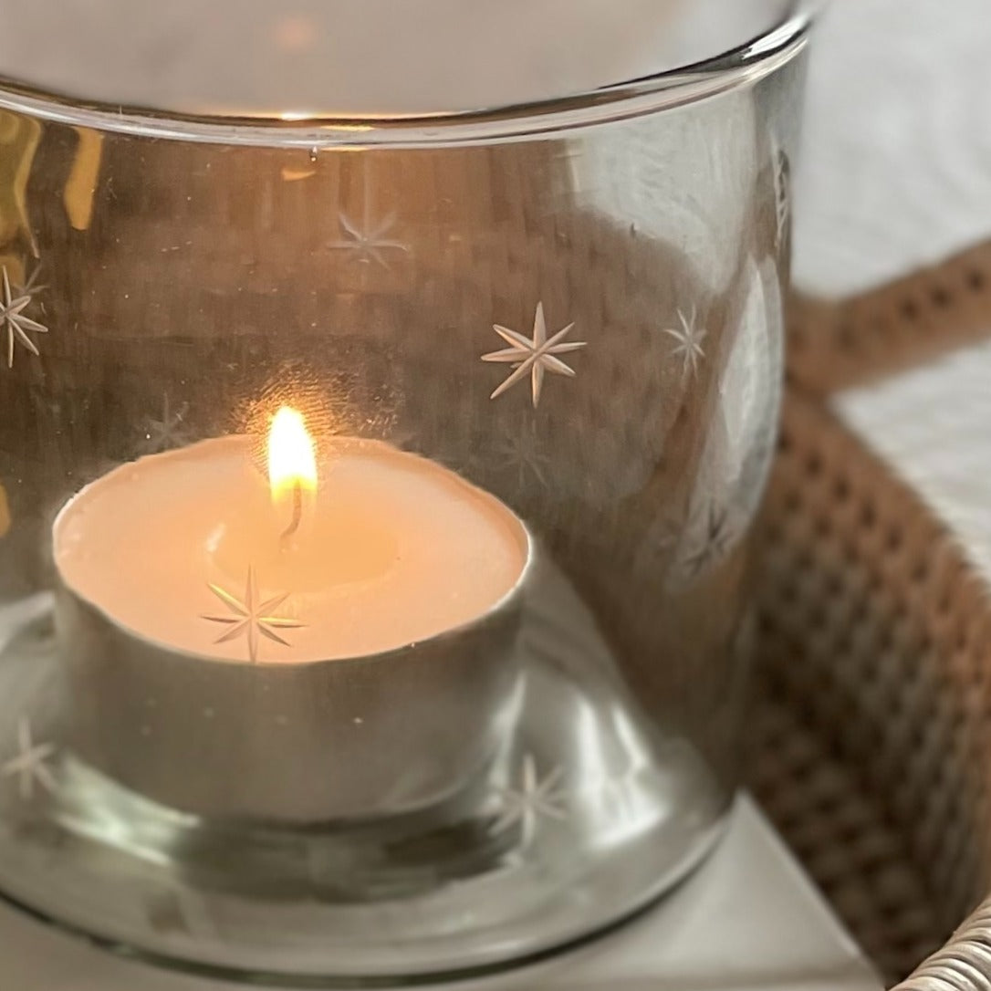 Glass hurricane candle holder with delicate etched star design, styled with a glowing candle on a round rattan tray.