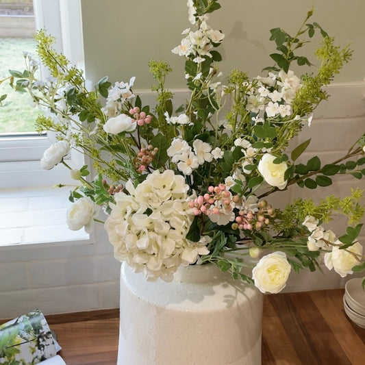 Faux spring bouquet with white ranunculus, wild roses, pink berries, mirabella sprays and a white hydrangea. Styled in a vase on a wooden kitchen worktop.