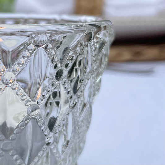 Close up of a clear glass tea light holder with an embossed diamond beaded pattern, styled on a table with soft, glowing candlelight.