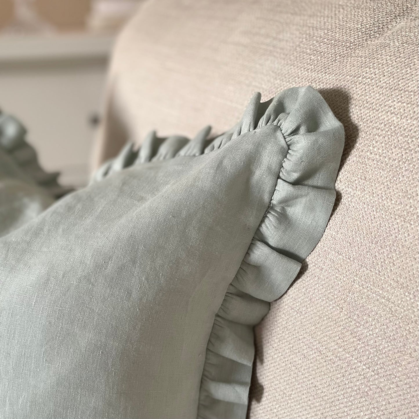 Close up of sage linen cushion with ruffled edge and mother of pearl button detail, styled on a neutral sofa.