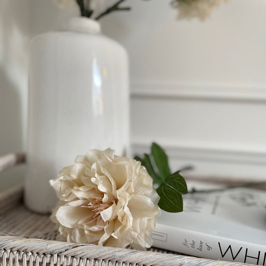 Soft faux peony stem with delicate cream/beige petals and green leaves styled in a vase on a rattan tray and book.