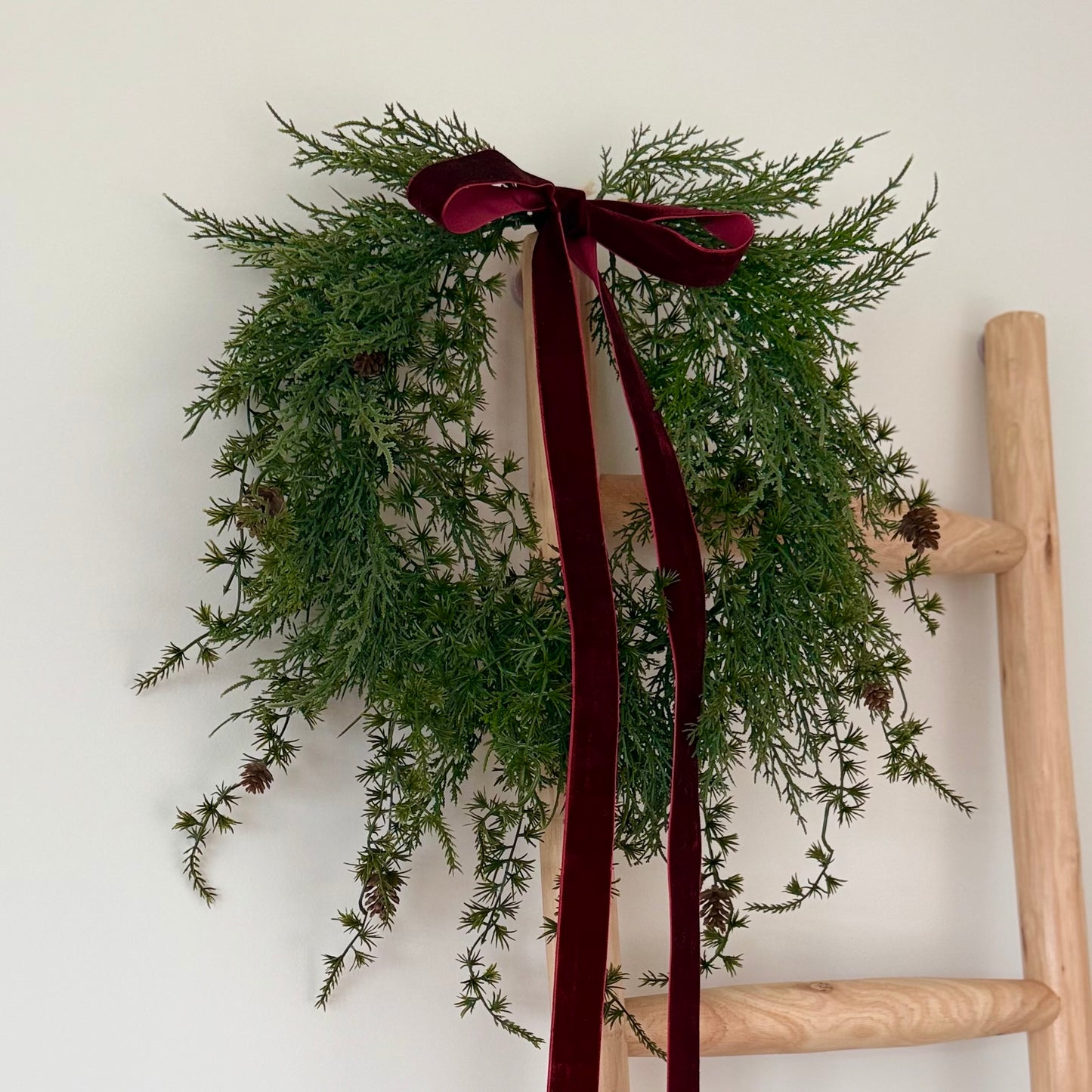 Green wreath with mini pinecones and a burgundy ribbon on a wooden ladder against a white wall
