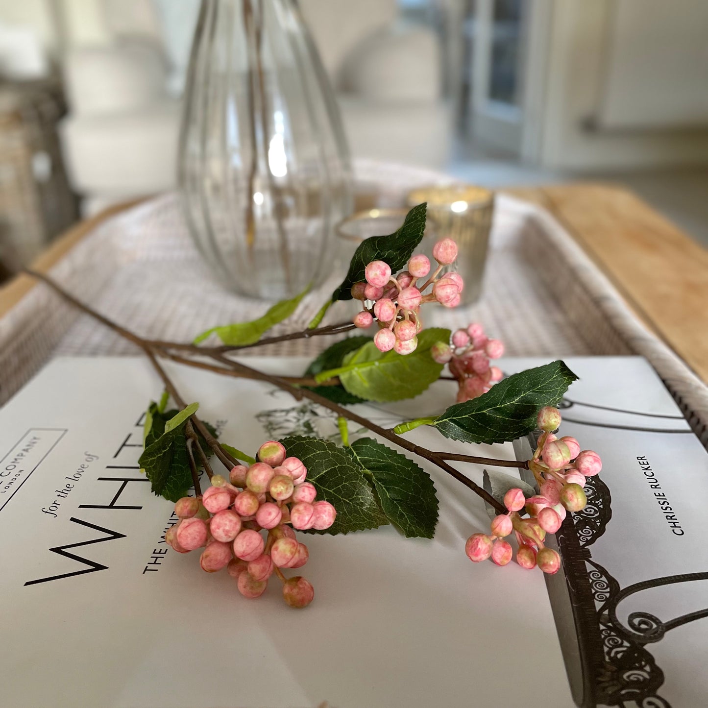 Faux pink berry stem with realistic clustered berries and green foliage, styled in a vase. On a rattan tray with a book.