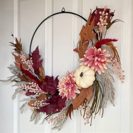Autumn hoop wreath with pampas grasses, maple leaves, pink berries, pink flowers and a neutral pumpkin. Styled on a white door.