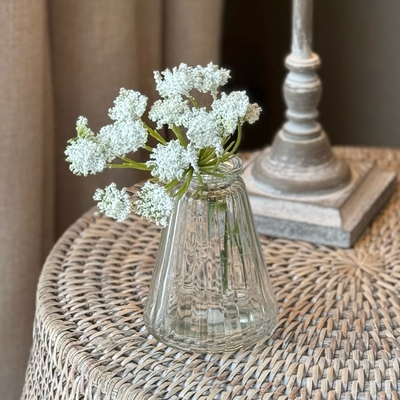 Clear glass bud vase with white flowers on a wicker surface.