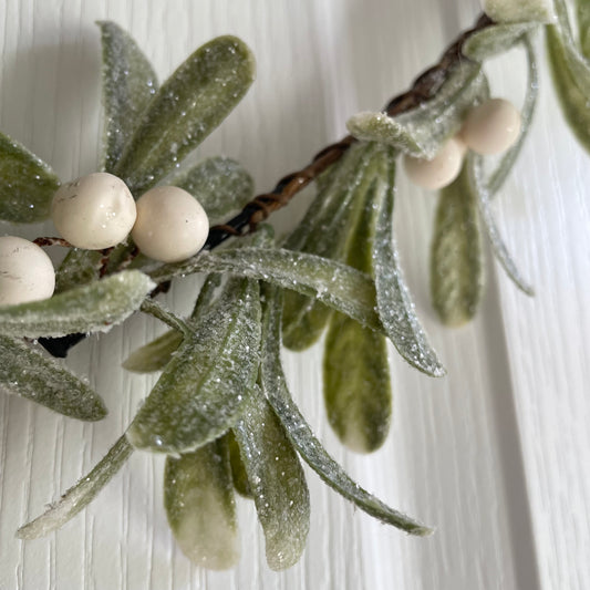 Heart shaped glittered mistletoe wreath with white berries, styled on a white door for a festive touch.