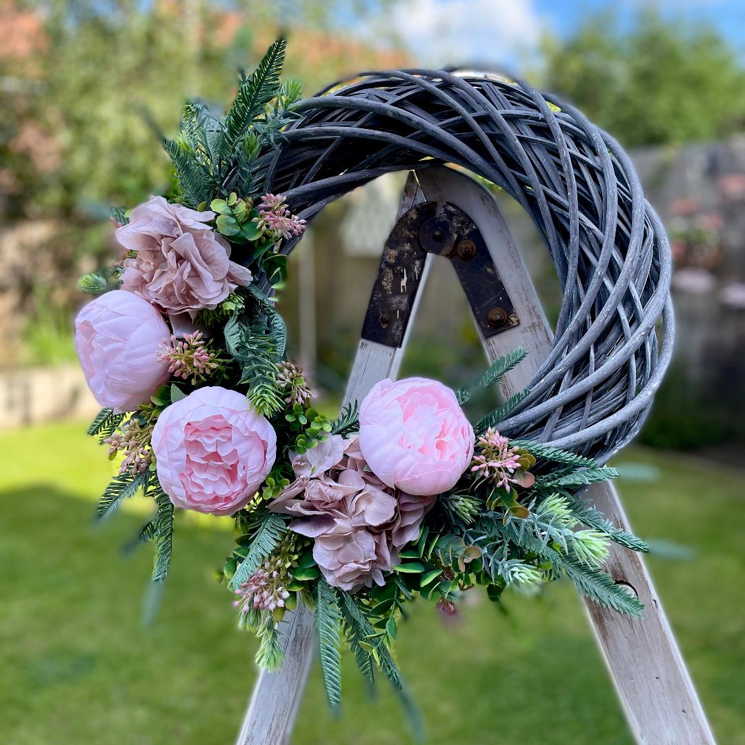 Pink peony and nude hydrangea wreath presented on a wooden ladder. Shown in a garden.