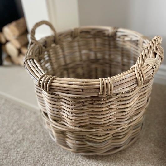Round rattan basket holding throws by a fireplace in a neutral, cosy interior.