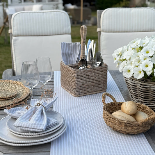 Whitewashed rattan caddy with handle for cutlery or condiments. Dining set up with plates, glasses, cutlery and bread on a tablecloth.