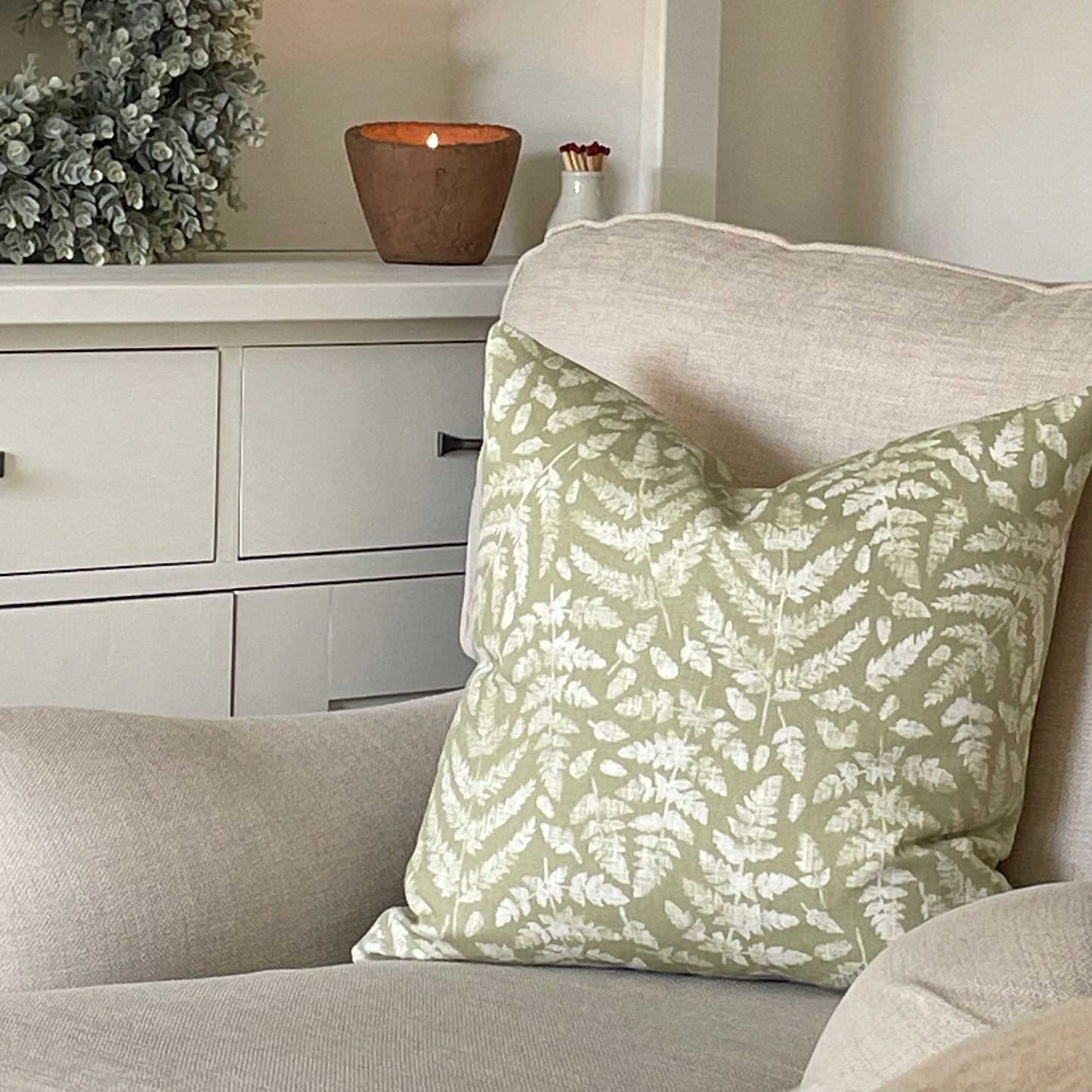 Sage green cushion with fern leaf detailing styled on a neutral armchair with a throw.