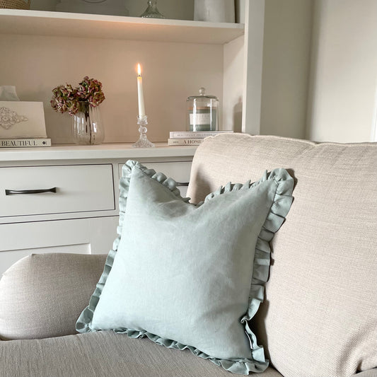 Sage linen cushion with ruffled edge and mother of pearl button detail, styled on a sofa. With white cabinet and candle styled in the background.