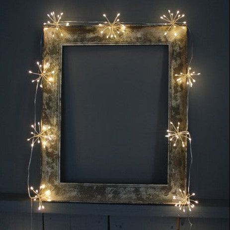 Warm white starburst string lights draped along a rustic picture frame, creating a soft festive glow indoors.