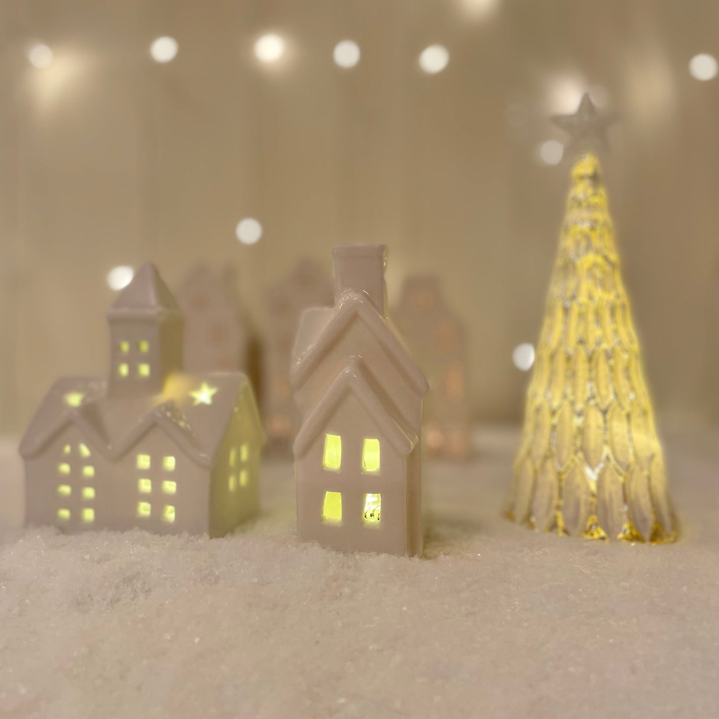 Small white glazed ceramic LED house ornament with warm light, styled on a snowy scene with other ceramic christmas decor.