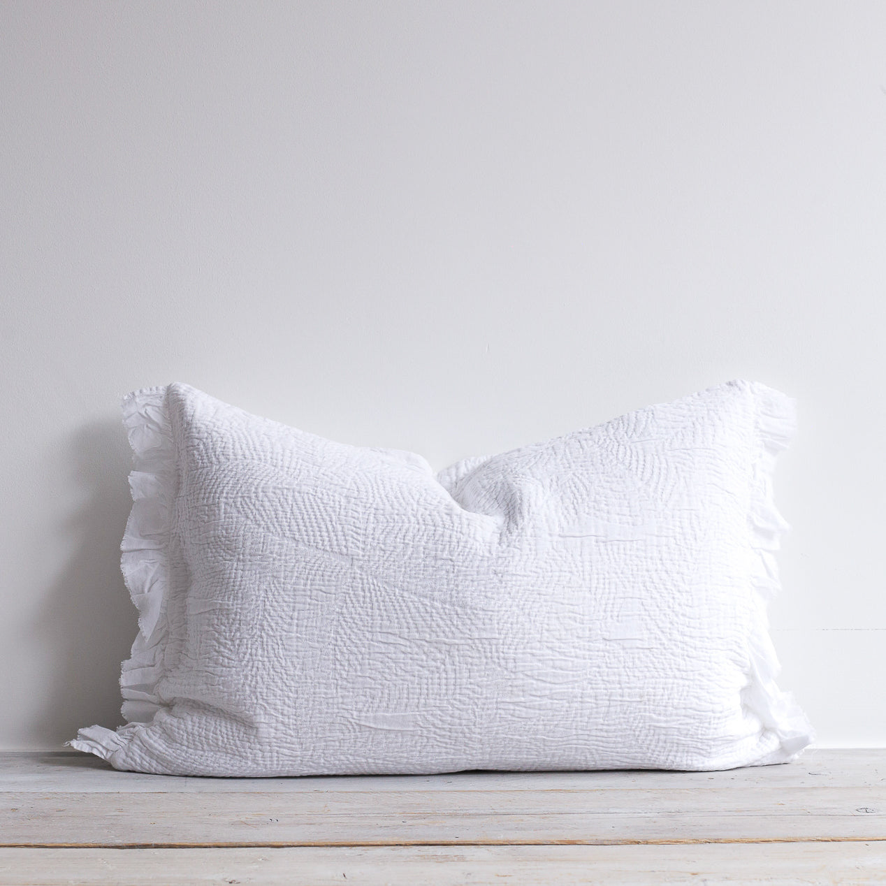 White quilted cushion with frilled edges and embroidered textured design. Styled on a wooden bench.