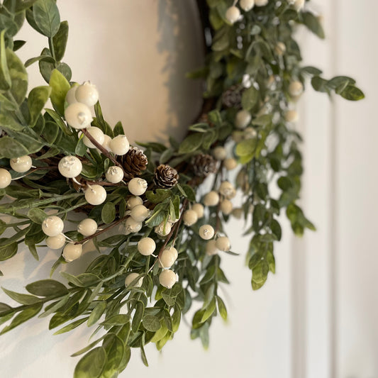 Winter wreath with faux green foliage, white berries and miniature pinecones on a grapevine base, styled on a wall.