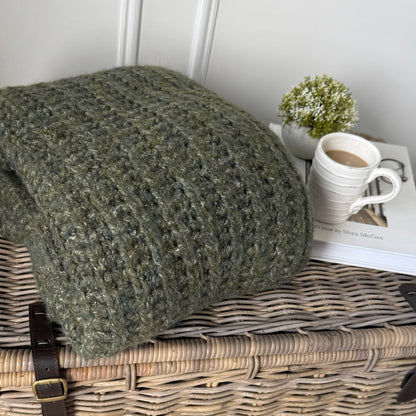 Green textured blanket on a wicker surface with a cup of coffee and plant in the background.