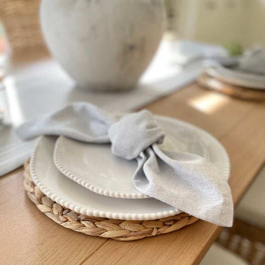 White porcelain side plate with organic shape and beaded detailing around the rim, styled on a wooden table with a grey napkin and seagrass place mat.