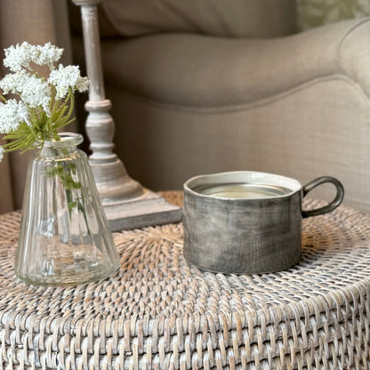 Decorative black wash ceramic mug style candle holder on a rattan side table with vase of flowers.