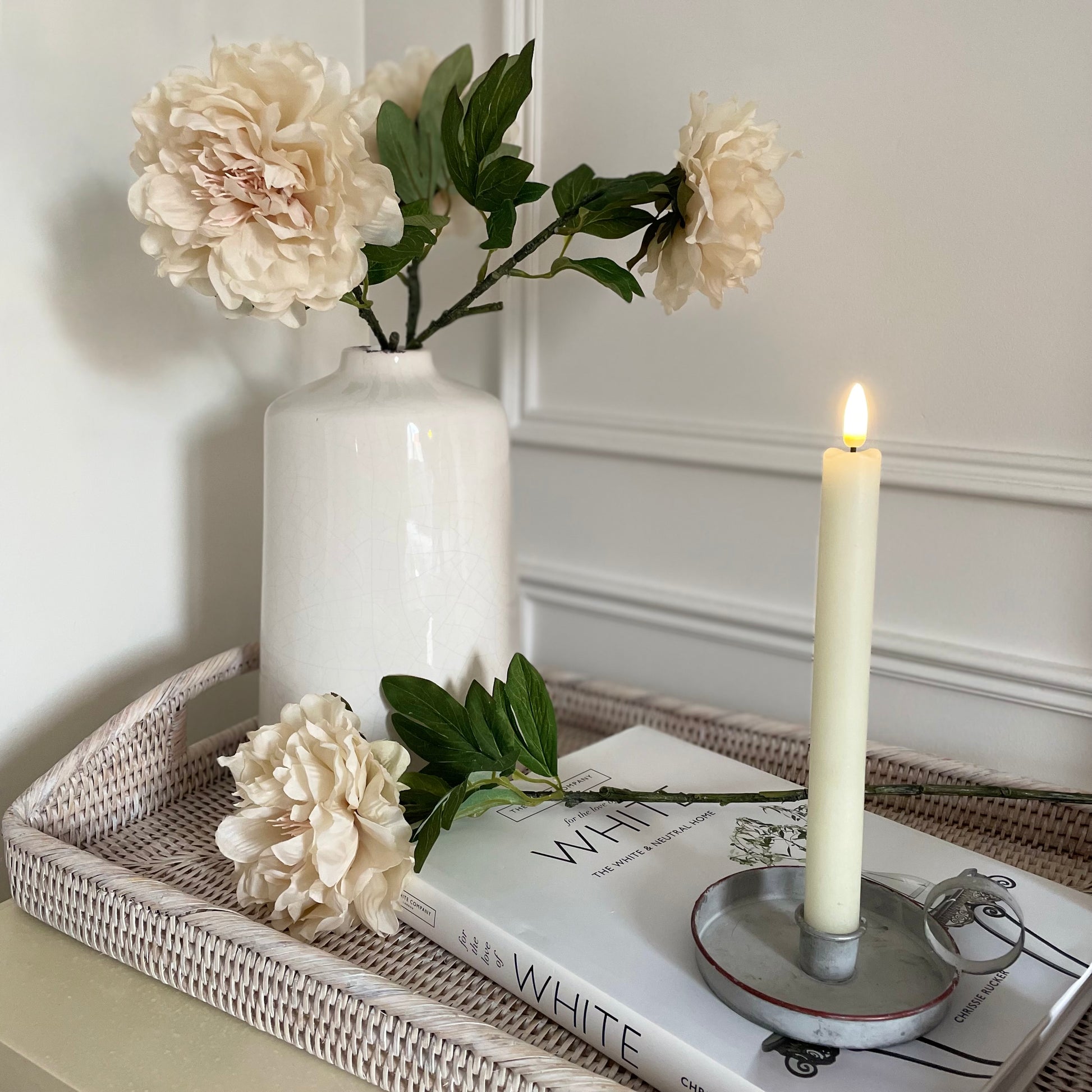 Soft faux peony stem with delicate cream petals and green leaves styled in a vase. On a rattan tray with a book and candle.
