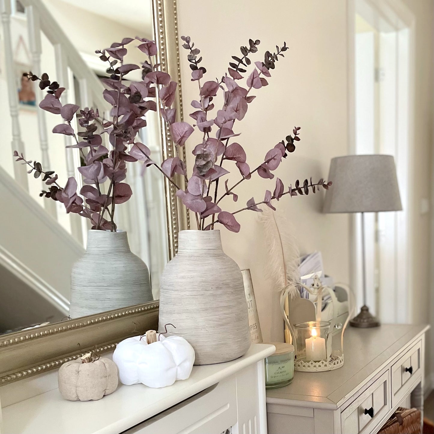Burgundy faux eucalyptus spray with realistic brown and green tones styled in a rustic ceramic vase. With decorative pumpkins and a lamp.