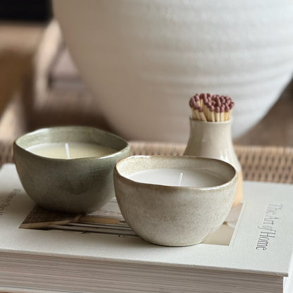 Two ceramic bowls with candles on a surface, with a vase and matchbox in the background.