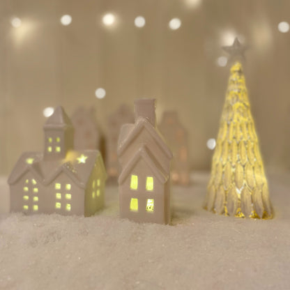 Small white ceramic church ornament with pointed steeple and warm LED light, styled on a snow scene with other christmas ceramic decor.