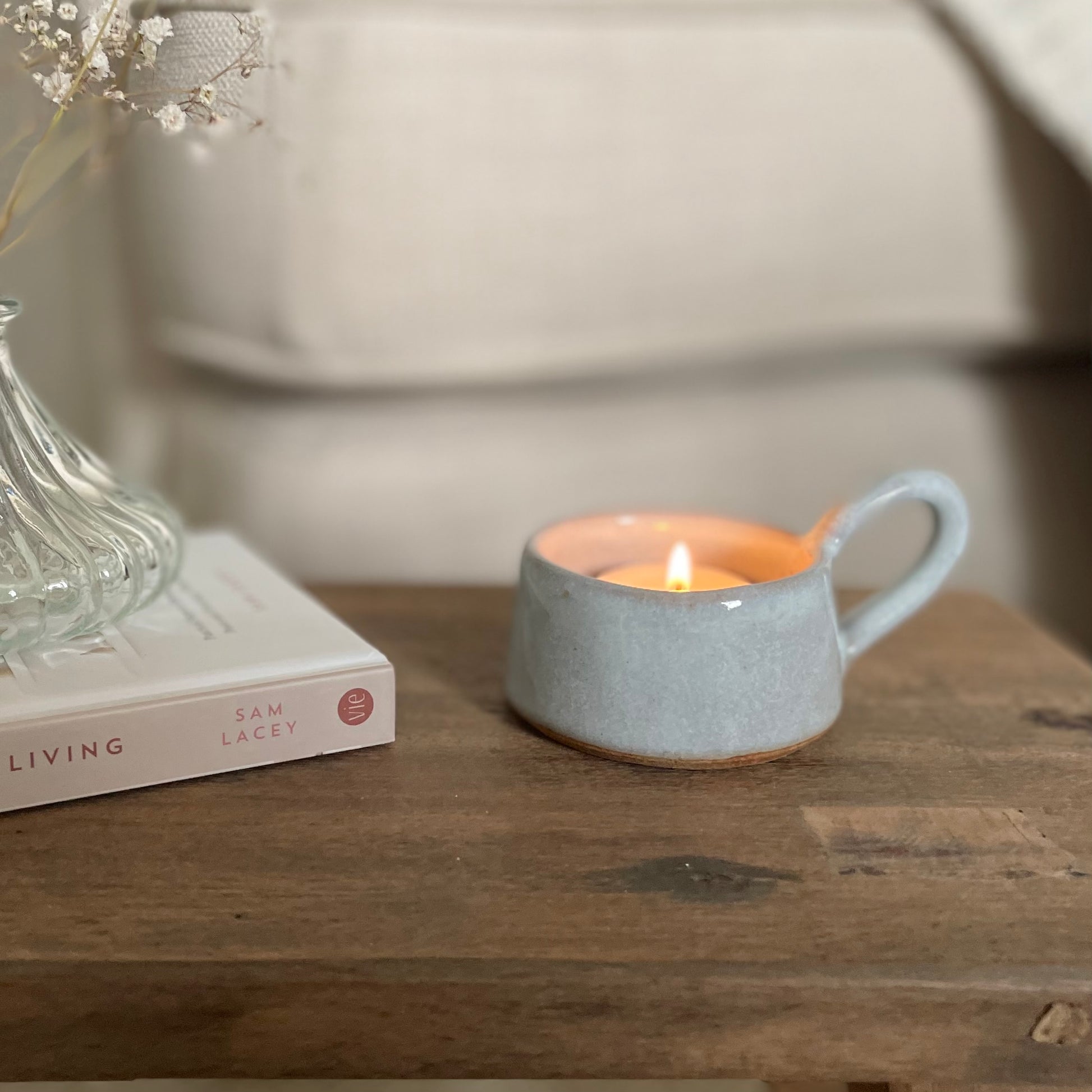 Eggshell blue ceramic tea light holder with handle, styled on a wooden stool with a book and cosy sofa.