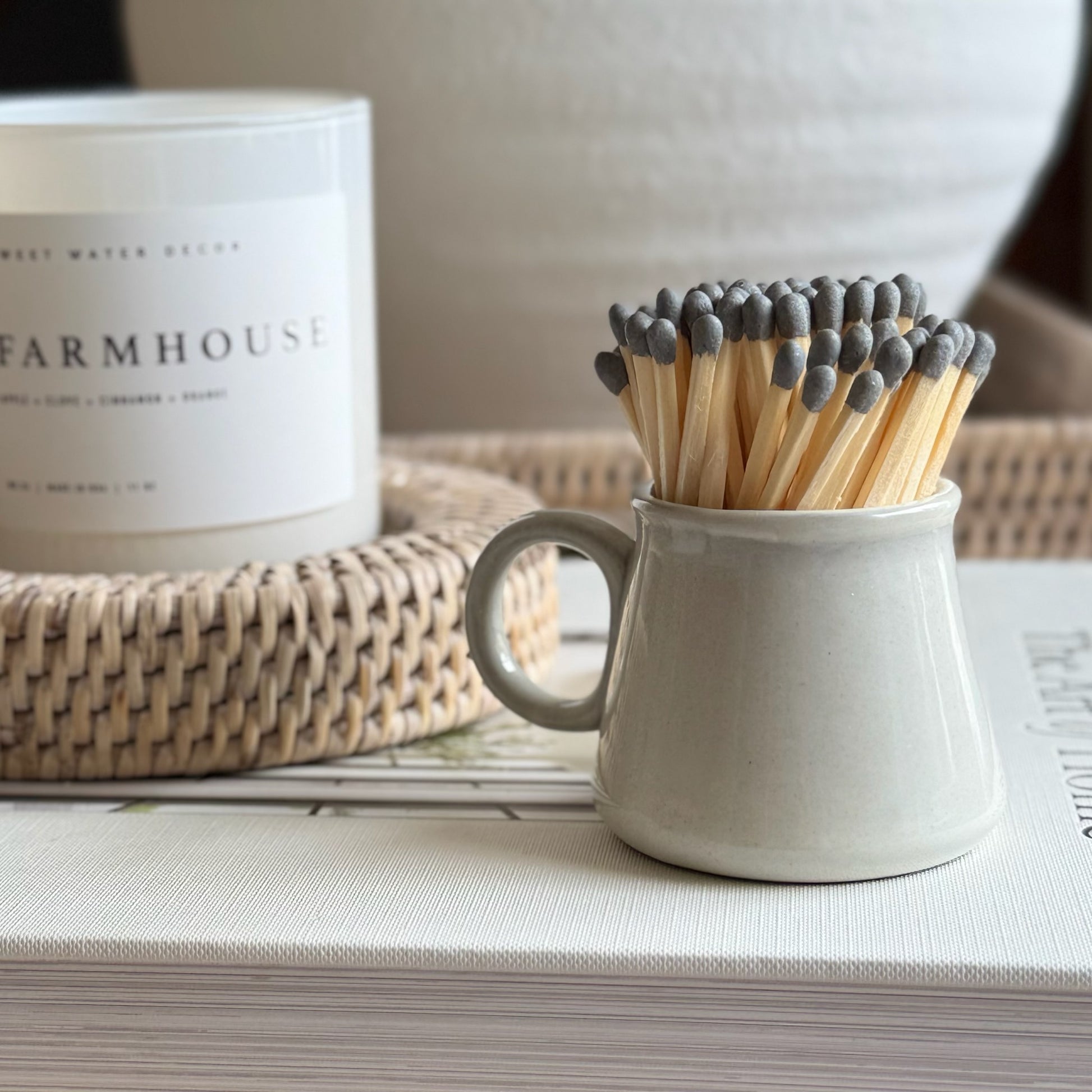 Matches in a ceramic cup on a surface with a candle labeled 'Farmhouse' in the background.