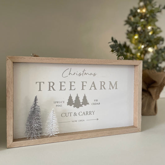 Wooden Christmas plaque with “Christmas Tree Farm” text, snowy bristle trees, and jute hanger in neutral tones. Styled on a white shelf with a lit up Christmas tree.