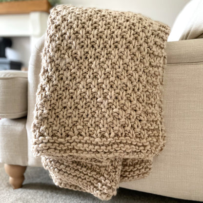 Chunky moss knitted oatmeal throw draped over a cream armchair in a cosy living room.