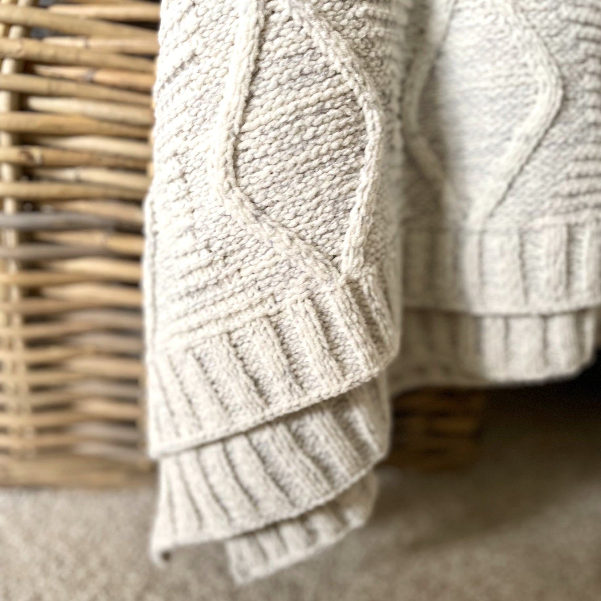 Soft melange grey chunky cable knit throw with ribbed edge draped over a rustic rattan basket.