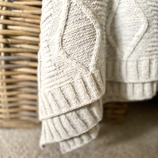 Soft melange grey chunky cable knit throw with ribbed edge draped over a rustic rattan basket.