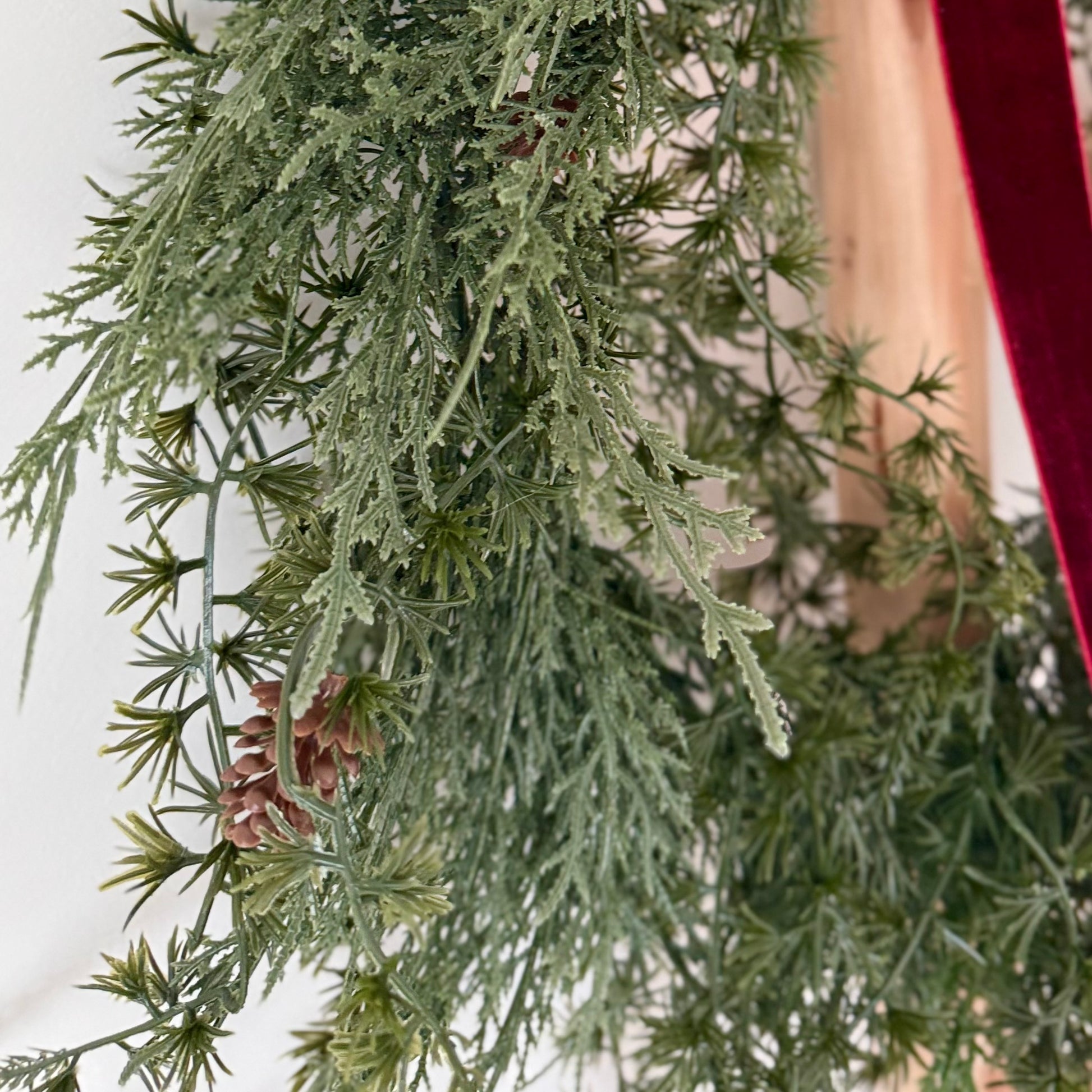 Close up of a green artificial wreath with pine cones and red velvet ribbon on a white background