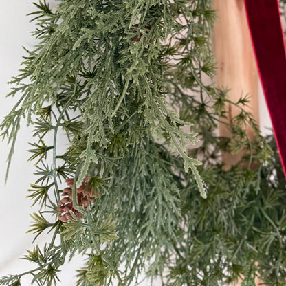 Close up of a green artificial wreath with pine cones and red velvet ribbon on a white background
