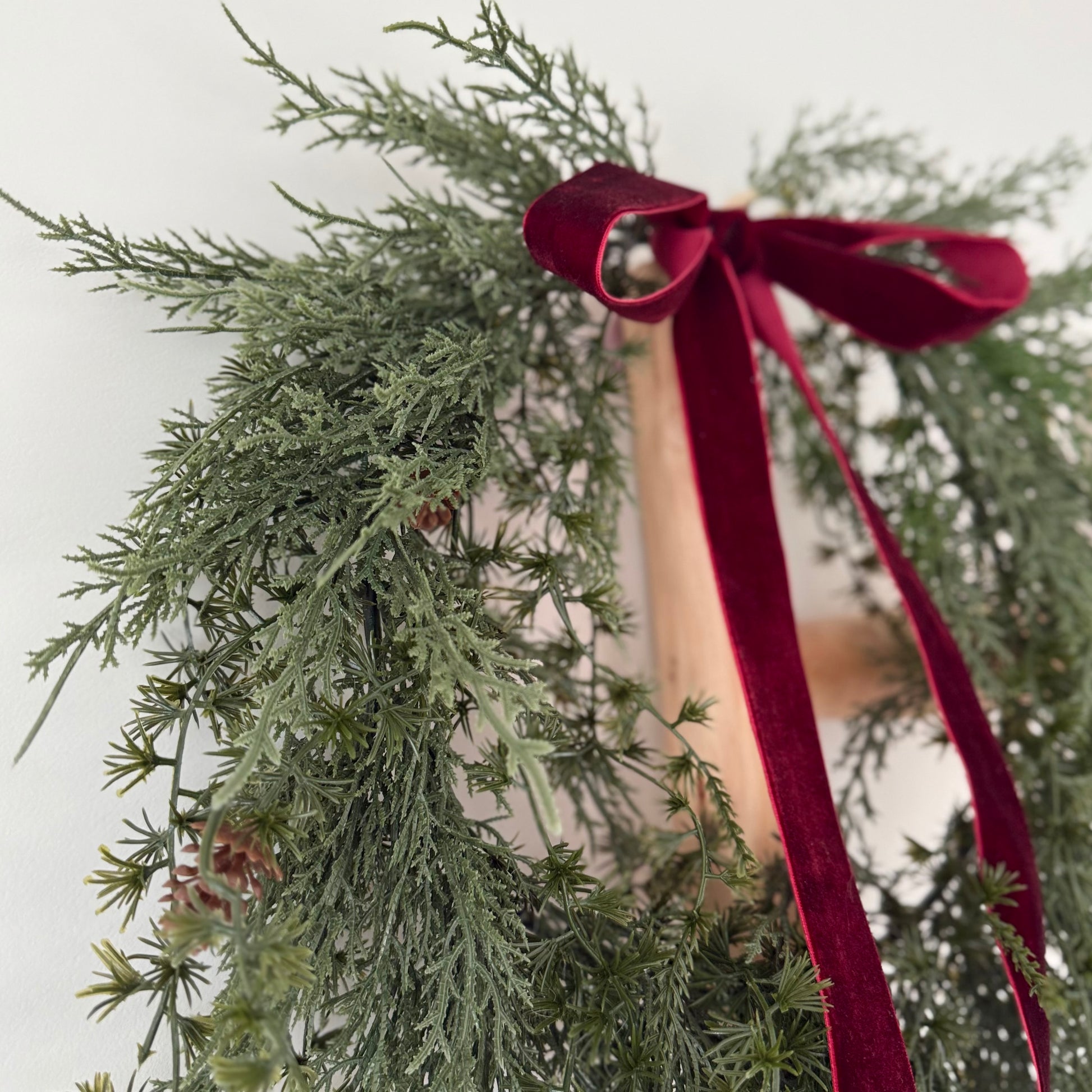 Green faux foliage wreath with mini pinecones with a red ribbon against a light background.