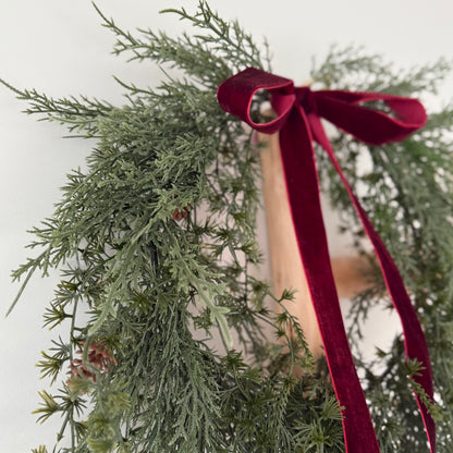 Green faux foliage wreath with mini pinecones with a red ribbon against a light background.