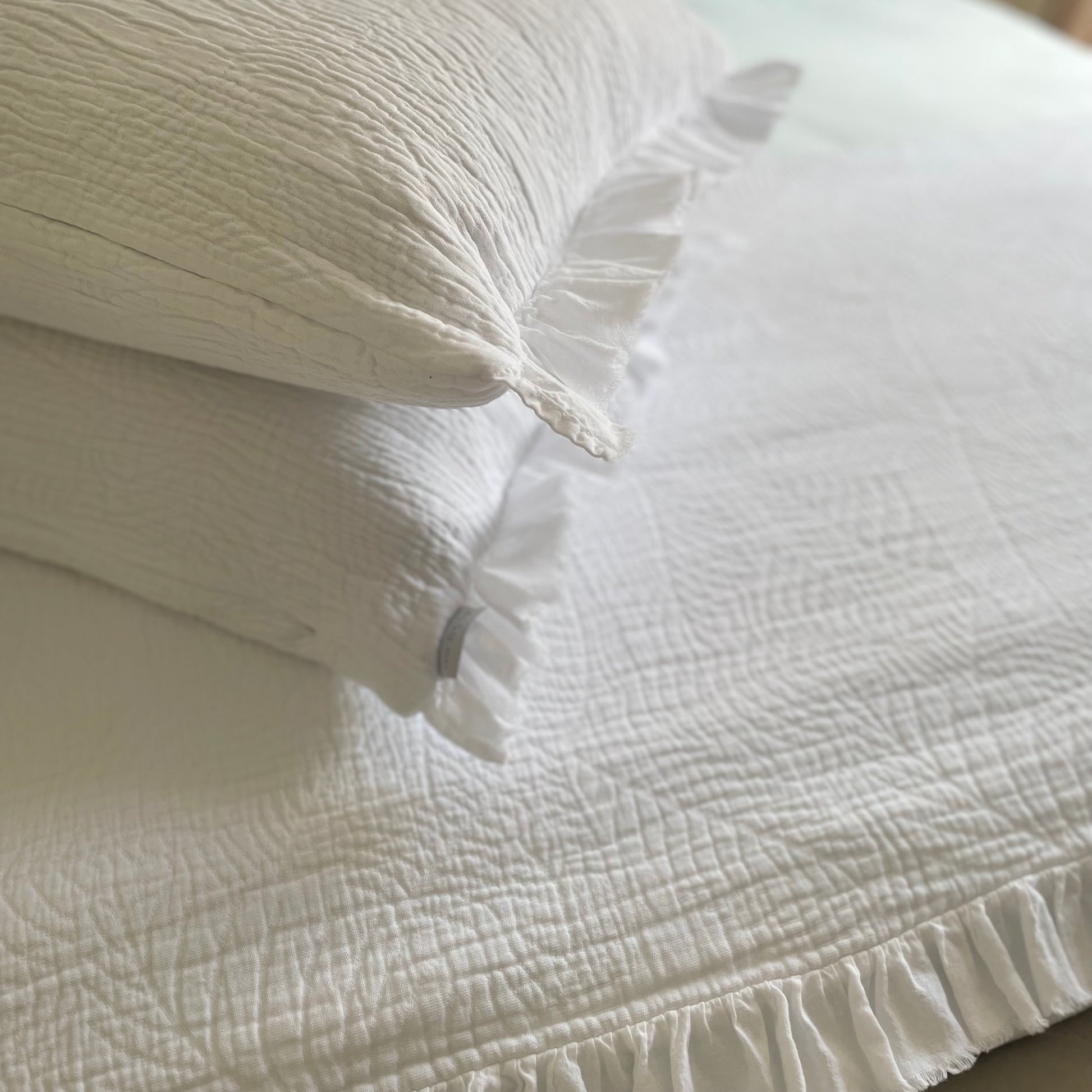 Close up of white quilted cushion with frilled edges and embroidered textured design. Styled on a bed.