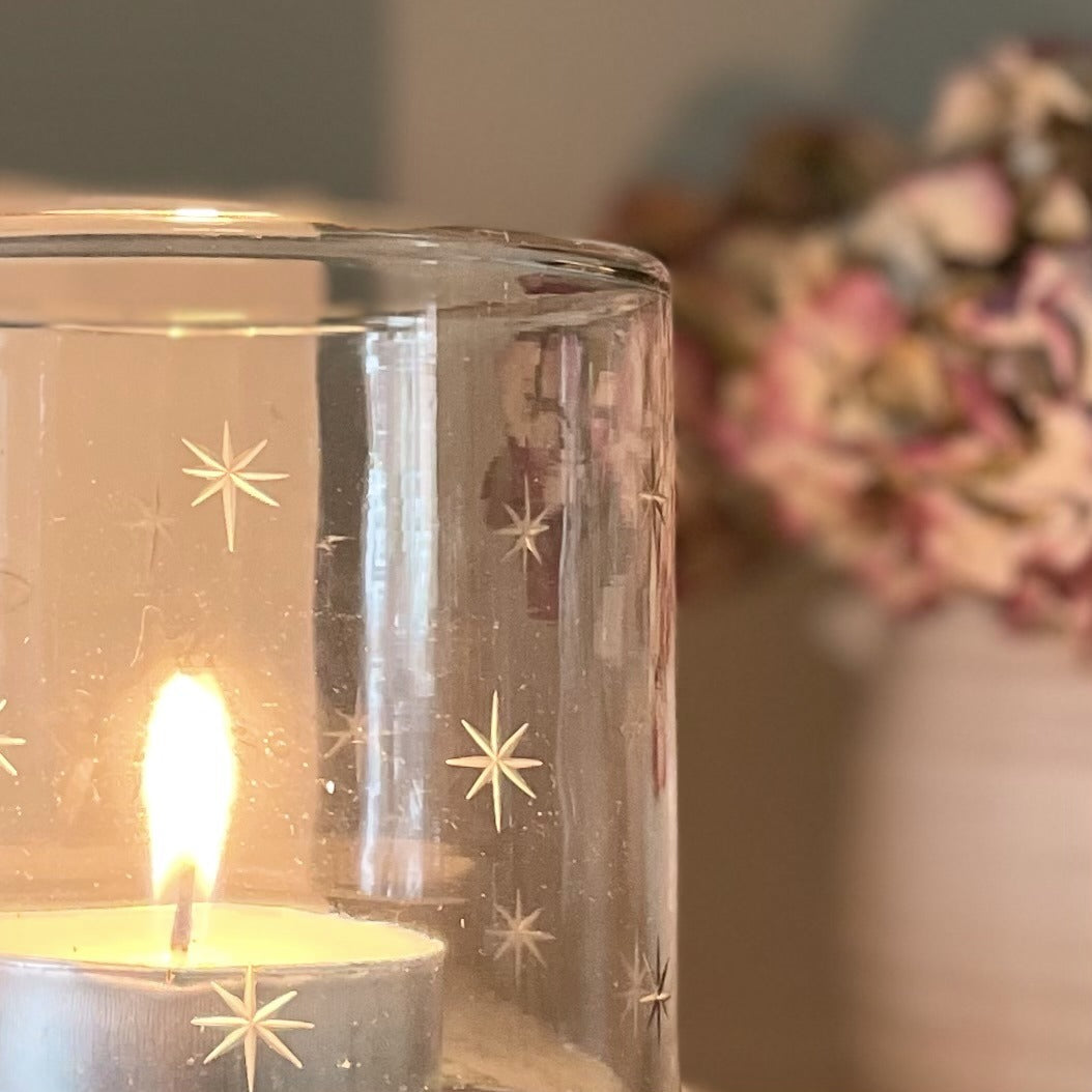 Glass hurricane candle holder with delicate etched star design, styled with a glowing candle and flowers behind.