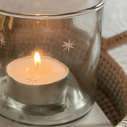 Glass hurricane candle holder with delicate etched star design, styled with a glowing candle on a round rattan tray.