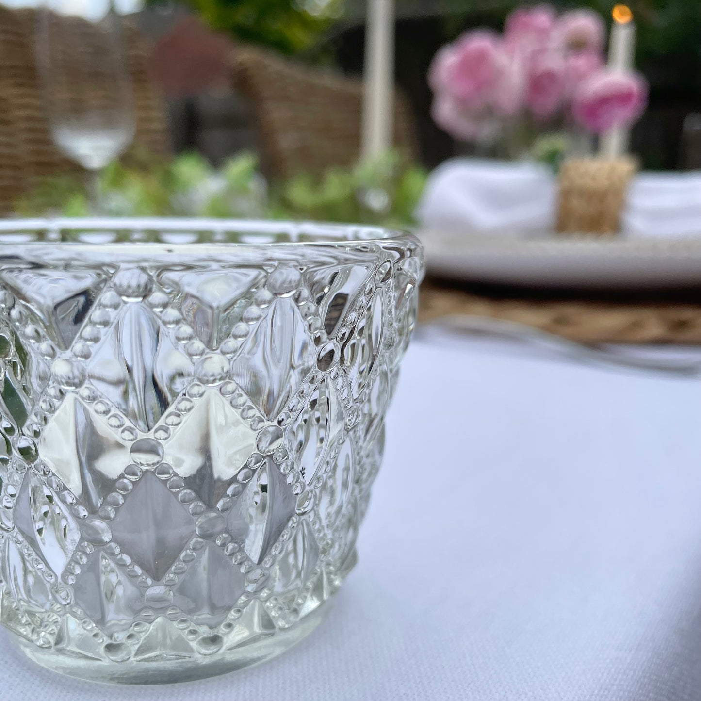 Clear glass tea light holder with an embossed diamond beaded pattern, styled on a table with soft, glowing candlelight. On a dining table in a rose garden.