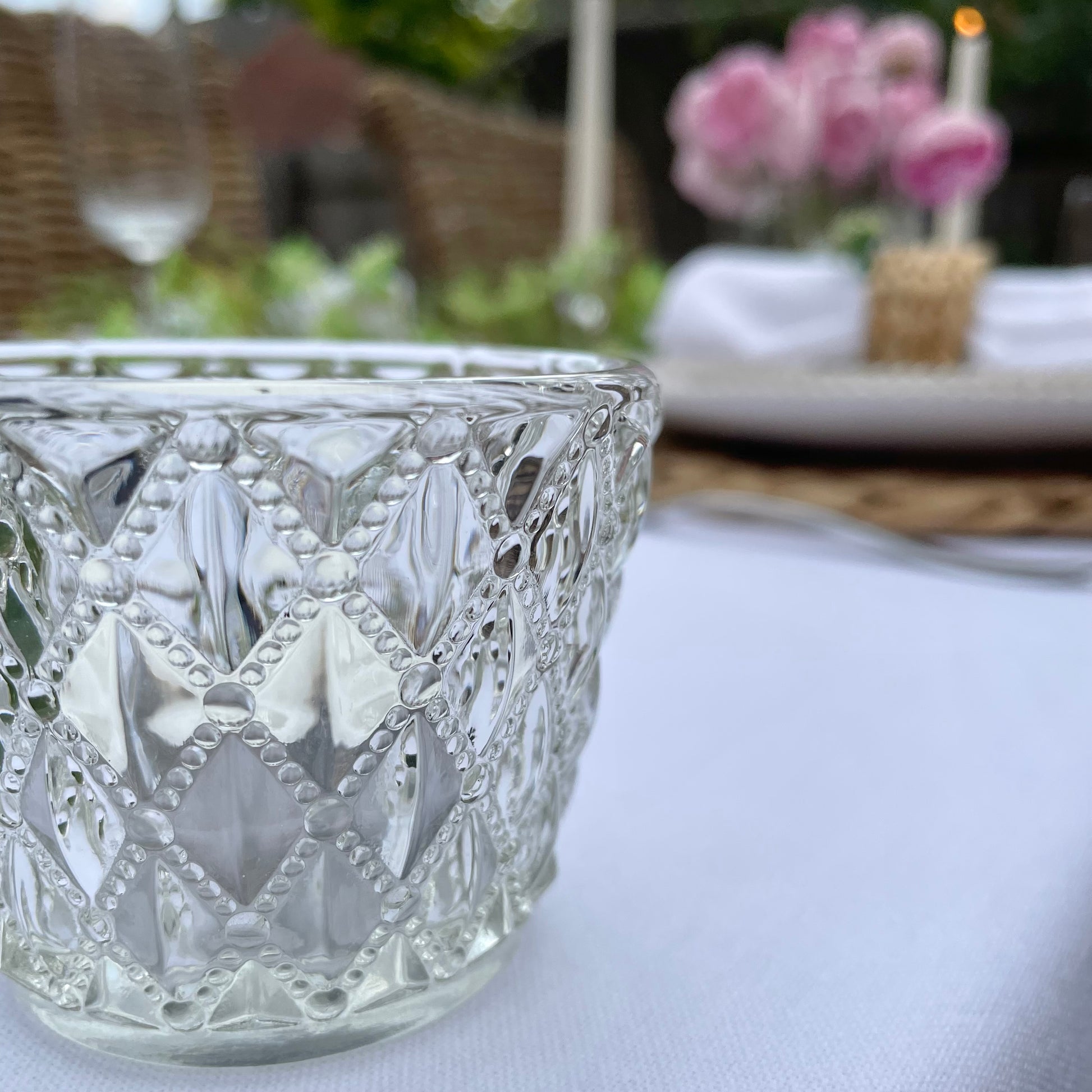 Clear glass tea light holder with an embossed diamond beaded pattern, styled on a table with soft, glowing candlelight. On a dining table in a rose garden.