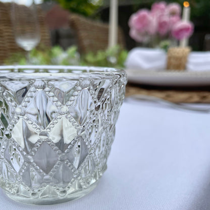 Clear glass tea light holder with an embossed diamond beaded pattern, styled on a table with soft, glowing candlelight. On a dining table in a rose garden.