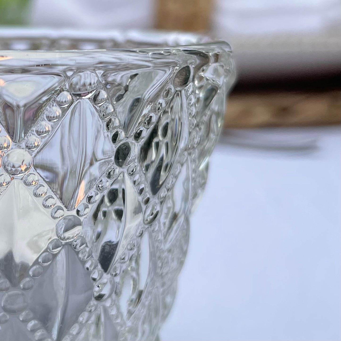 Close up of a clear glass tea light holder with an embossed diamond beaded pattern, styled on a table with soft, glowing candlelight.