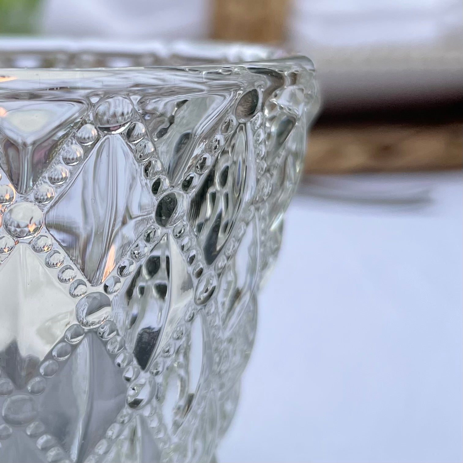 Close up of a clear glass tea light holder with an embossed diamond beaded pattern, styled on a table with soft, glowing candlelight.