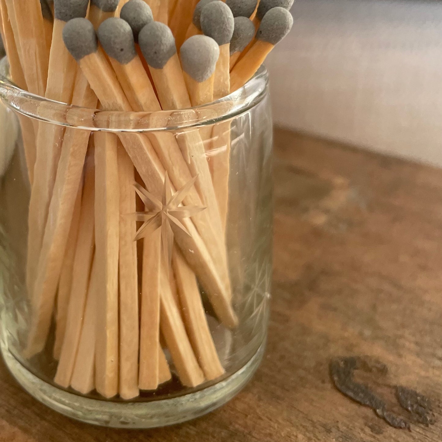 Glass match pot with etched star detail, grey matches, and hidden striker pad on the base. Styled on a rustic wooden stool.