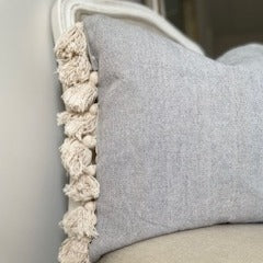 Grey textured cotton cushion with cream tassels on each edge, styled on a neutral chair in a cosy hall setting.