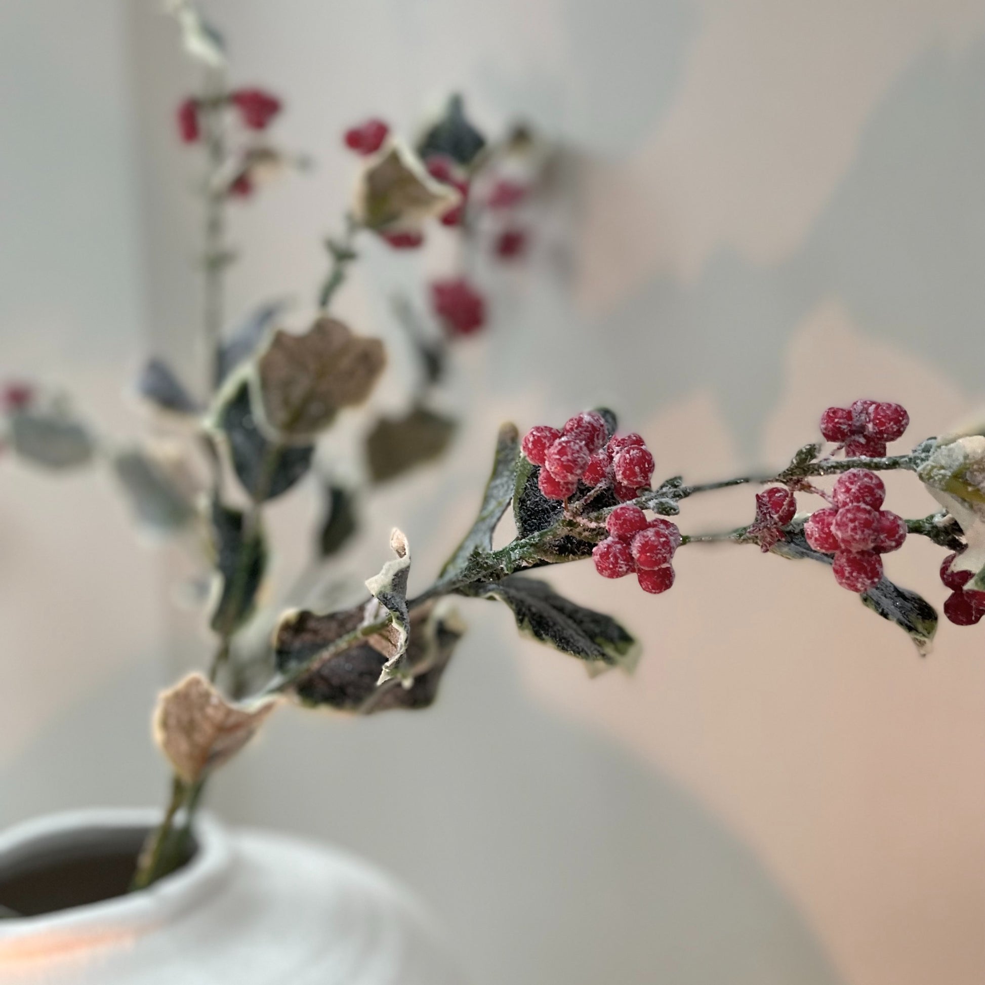Frosted holly stem with red berries styled in stone vase, festive faux foliage 68 cm. Placed on white kitchen worktop.